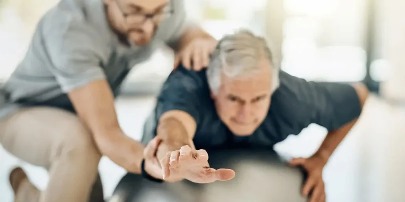 Physiotherapeut unterstützt älteren Mann bei einer Gleichgewichts- und Dehnübung auf einem Gymnastikball zur Verbesserung der Mobilität und Rehabilitation.