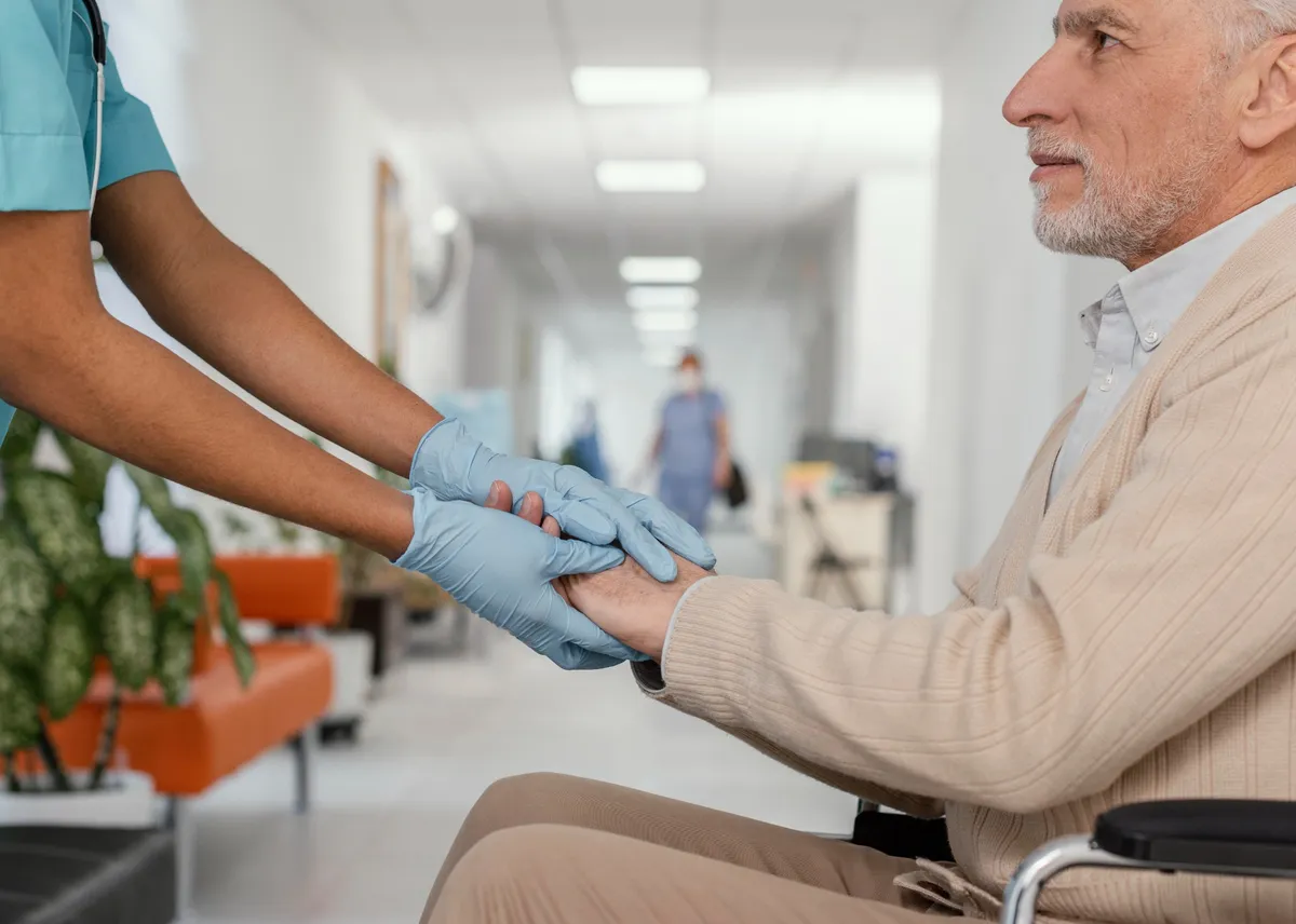 Pflegekraft mit Handschuhen hält unterstützend die Hand eines älteren Mannes im Rollstuhl im Krankenhausflur.