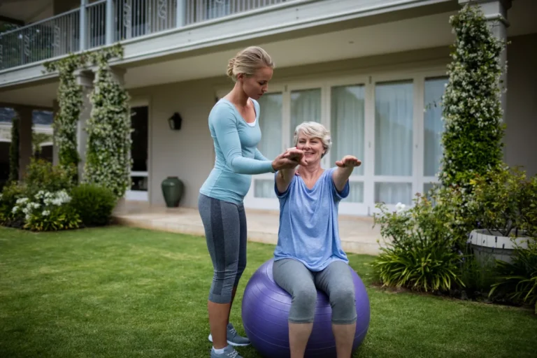 Fitnesstrainerin hilft einer älteren Frau bei Gymnastikübungen im Garten auf einem Gymnastikball.