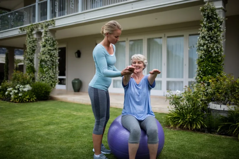 female-coach-assisting-senior-woman-performing-exercise_1200 Fitnesstrainerin hilft einer älteren Frau bei Gymnastikübungen im Garten auf einem Gymnastikball.