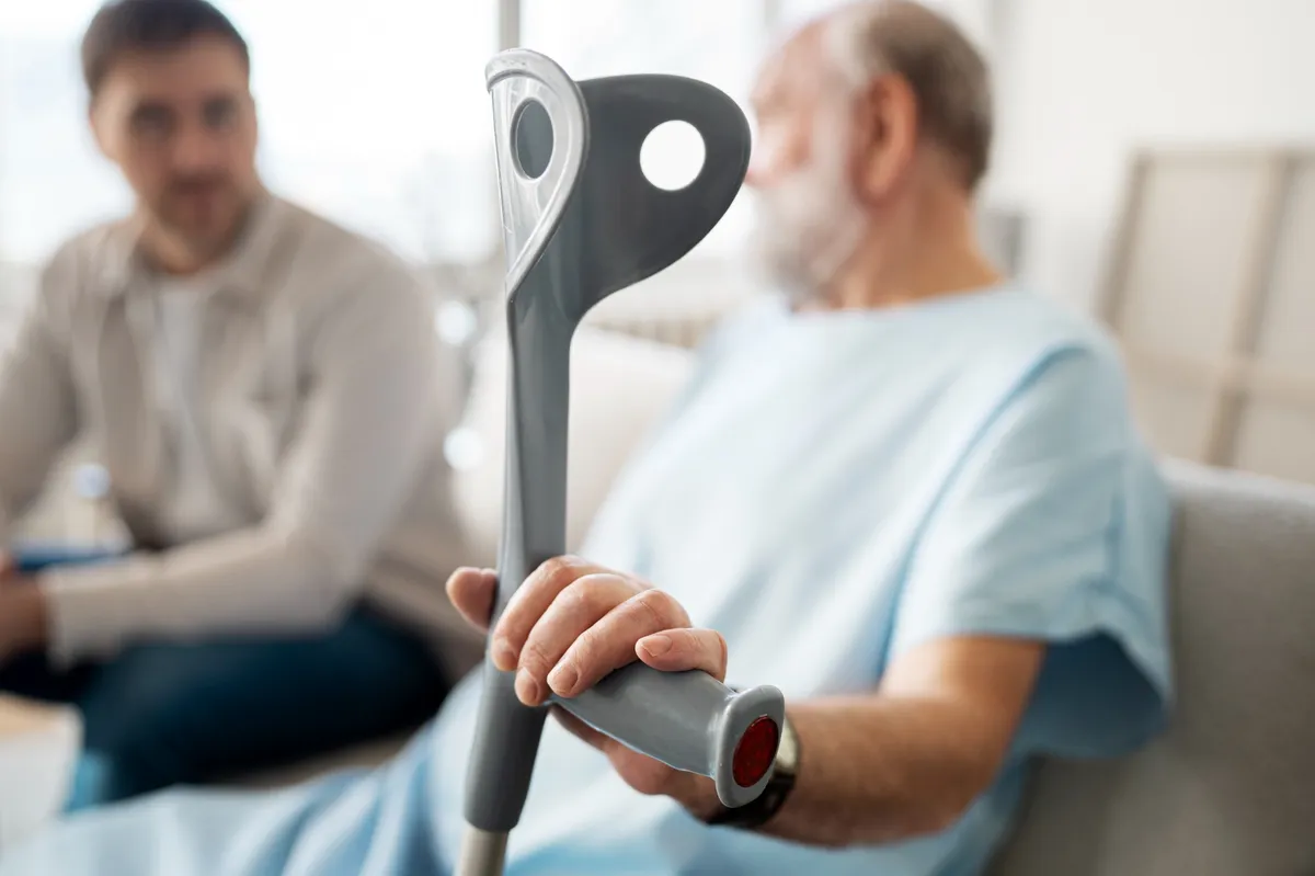 Älterer Patient in Klinikbekleidung hält eine Gehhilfe, im Hintergrund sitzt eine begleitende Person.