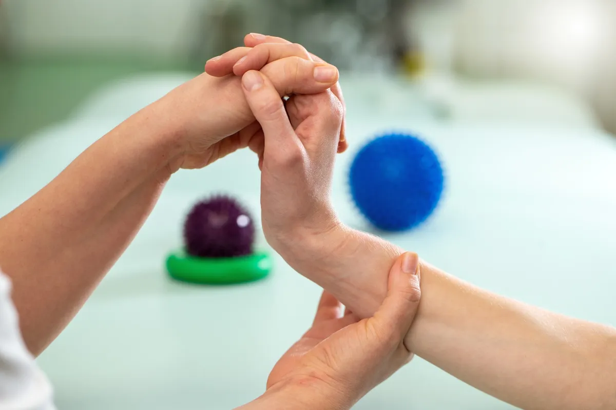 Therapeutin führt Mobilisationsübung am Handgelenk einer Patientin durch, im Hintergrund liegen zwei Igelbälle auf einer Behandlungsliege.