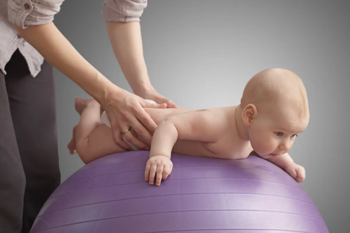 Baby liegt bäuchlings auf einem lila Gymnastikball, während eine Frau sanft den Rücken massiert – therapeutische Übung zur Förderung der motorischen Entwicklung.