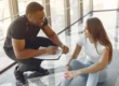 Ein Fitnesstrainer spricht mit einer jungen Frau im Trainingsoutfit, die auf einer Matte sitzt – beide lächeln und wirken motiviert.
