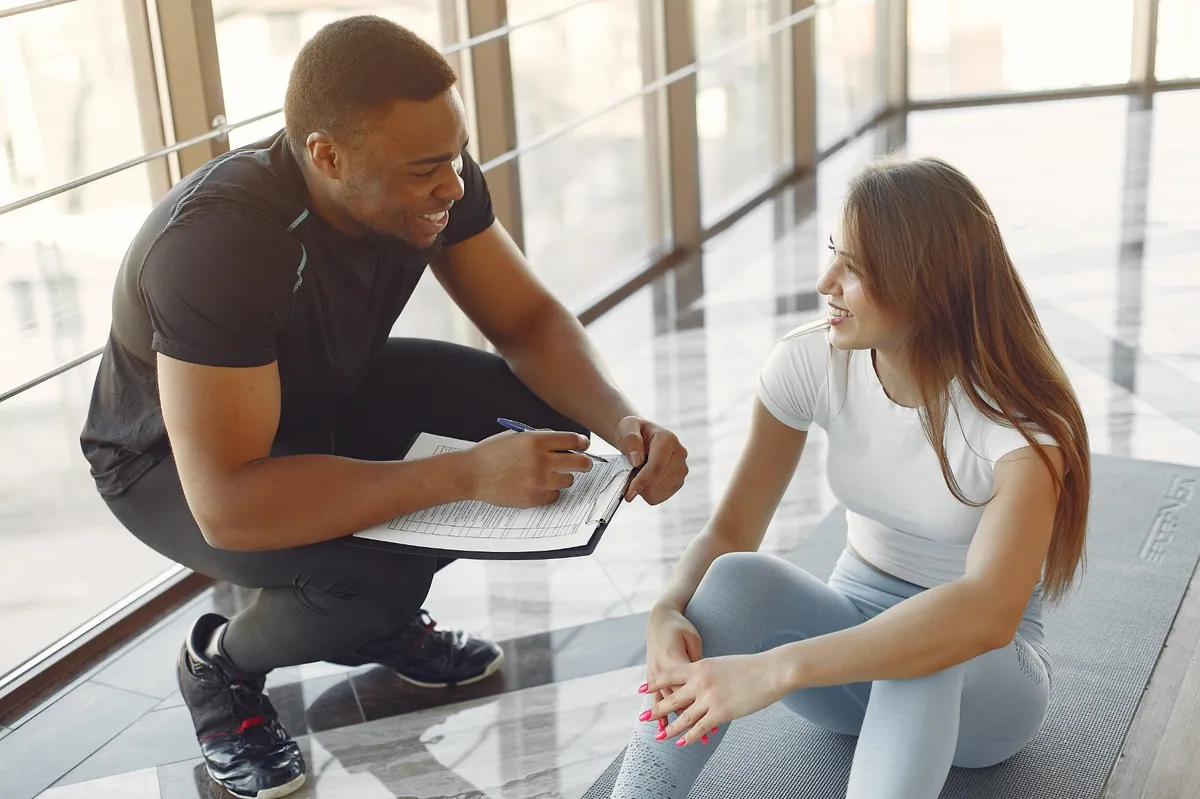 Ein Fitnesstrainer spricht mit einer jungen Frau im Trainingsoutfit, die auf einer Matte sitzt – beide lächeln und wirken motiviert.