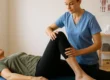 Eine Physiotherapeutin in blauer Kleidung führt eine Mobilisationsübung am Knie einer älteren Frau durch, die entspannt auf einer Behandlungsliege liegt. Der Raum wirkt hell und professionell.