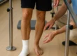 Ein junger Mann mit bandagiertem Fuß trainiert an einer Gehhilfe in einer Reha-Einrichtung. Ein Therapeut kniet neben ihm und gibt unterstützende Anweisungen.