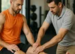 Ein Physiotherapeut tastet das Knie eines sportlich gekleideten Mannes ab, der auf einer Behandlungsliege sitzt. Die Szene spielt sich in einem Trainingsraum mit Fitnessgeräten im Hintergrund ab.