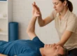 Physiotherapeutin führt Mobilisationsübung mit dem rechten Arm eines liegenden älteren Mannes durch – Fokus auf Schultergelenk und Beweglichkeit.