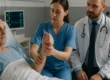 Eine Physiotherapeutin überprüft die Beweglichkeit des Arms eines älteren Mannes, der in einem Krankenhausbett liegt, während ein Arzt daneben sitzt.